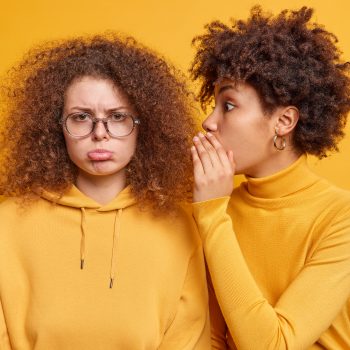 Surprised African American woman whispers secret information on ear of best friend who looks with gloomy expression spread rumors tells private news isolated over yellow background. Secrecy concept
