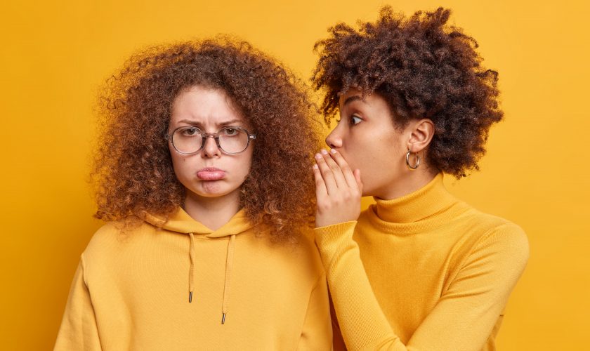 Surprised African American woman whispers secret information on ear of best friend who looks with gloomy expression spread rumors tells private news isolated over yellow background. Secrecy concept